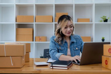 A beautiful Asian business owner opens an online store, she is checking orders from customers via laptop, sending goods through a courier company, concept of a woman opening an online business.