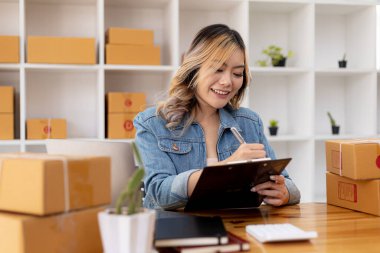 Bir e-ticaret şirketini işleten kadın, onlara göndermeden önce müşterilerin bir listesini kağıda yazıyor. Web sitelerinde ve sosyal medyada bir e-ticaret şirketi işletiyor. Çevrimiçi ürün satma kavramı.