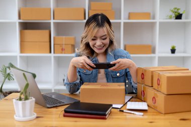 Paket kutularının önünde fotoğraf çekmek için akıllı telefon kullanan bir kadın. Malları paketlemek için paket kutuları, özel kurye şirketleri aracılığıyla mal teslimatı. Çevrimiçi satış ve online alışveriş kavramları.