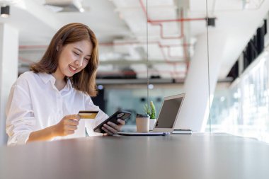 Tezgahta elinde cep telefonu ve kredi kartıyla oturan bir kadın, akıllı bir telefondan alışveriş yapıyor, mal ve hizmetleri kredi kartıyla ödüyor. Ödeme için kredi kartı kullanma kavramı.