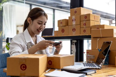 Asyalı bir kadın kanıt için paket kutusunun önünde fotoğraf çekmek için akıllı telefon kullanıyor, bir online mağazası var, özel bir nakliye şirketi aracılığıyla paketleyip gönderiyor. Çevrimiçi satış kavramı.