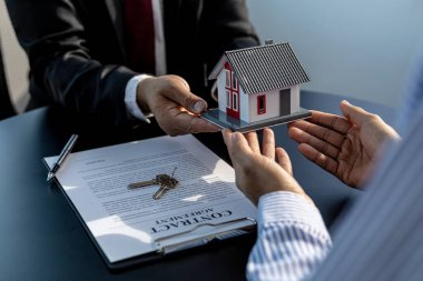 İki kişi küçük bir model ev tutuyor, ev satıcısı kontratı imzaladıktan ve evi inceledikten sonra evi müşteriye devrediyor. Gayrimenkul ticareti.