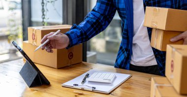 Paket kutuları tutan ve tabletten sipariş alan Asyalı bir adam, bir online mağazası var, özel bir nakliye şirketi aracılığıyla paketleyip gönderiyor. Çevrimiçi satış ve online alışveriş kavramları.