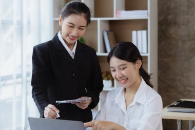 Girişim şirketinin atmosferinde, iki kadın çalışan, satışları artırmak ve yöneticilere raporlar hazırlamak için özetler ve pazarlama planları üzerinde çalışmak üzere fikir alışverişinde bulunuyorlar..