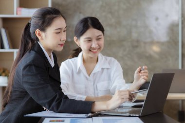 Girişim şirketinin atmosferinde, iki kadın çalışan, satışları artırmak ve yöneticilere raporlar hazırlamak için özetler ve pazarlama planları üzerinde çalışmak üzere fikir alışverişinde bulunuyorlar..