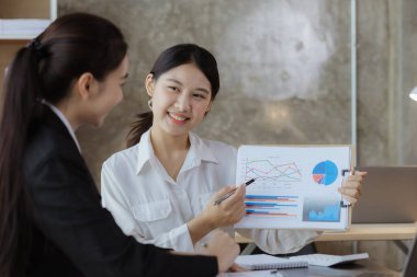 Girişim şirketinin atmosferinde, iki kadın çalışan, satışları artırmak ve yöneticilere raporlar hazırlamak için özetler ve pazarlama planları üzerinde çalışmak üzere fikir alışverişinde bulunuyorlar..