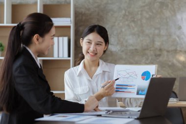 Girişim şirketinin atmosferinde, iki kadın çalışan, satışları artırmak ve yöneticilere raporlar hazırlamak için özetler ve pazarlama planları üzerinde çalışmak üzere fikir alışverişinde bulunuyorlar..