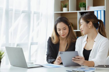 Girişim şirketinin atmosferinde, iki kadın çalışan, satışları artırmak ve yöneticilere raporlar hazırlamak için özetler ve pazarlama planları üzerinde çalışmak üzere fikir alışverişinde bulunuyorlar..