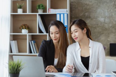 Girişim şirketinin atmosferinde, iki kadın çalışan, satışları artırmak ve yöneticilere raporlar hazırlamak için özetler ve pazarlama planları üzerinde çalışmak üzere fikir alışverişinde bulunuyorlar..