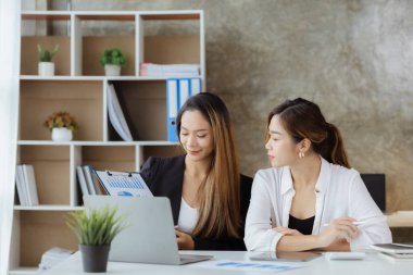 Girişim şirketinin atmosferinde, iki kadın çalışan, satışları artırmak ve yöneticilere raporlar hazırlamak için özetler ve pazarlama planları üzerinde çalışmak üzere fikir alışverişinde bulunuyorlar..