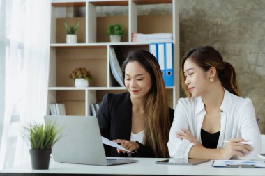 Girişim şirketinin atmosferinde, iki kadın çalışan, satışları artırmak ve yöneticilere raporlar hazırlamak için özetler ve pazarlama planları üzerinde çalışmak üzere fikir alışverişinde bulunuyorlar..