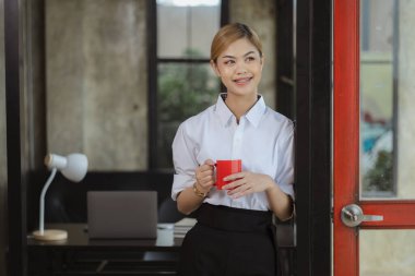 Asian female employee in company office standing holding red coffee cup, energetic young Asian businesswoman founded a new modern management startup company. female leaders running a business Concept.