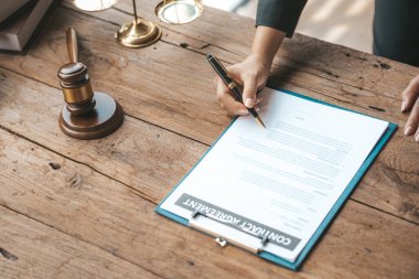 A lawyer sits in his office, on a table with a small hammer to beat the judges desk in court. and justice scales, lawyers are drafting a contract for the client to use with the defendant to sign.