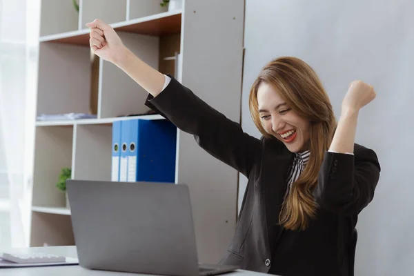 An Asian female employee is in a company office, looking at data from a laptop and showing joy, she is an employee of a startup marketing department. Company sales management concept.