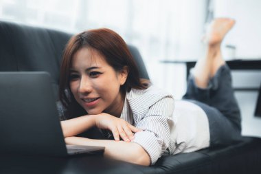 Asian woman lying on the sofa playing laptop on the sofa, she is taking a weekend break after a hard day's work. Holiday relaxation ideas.