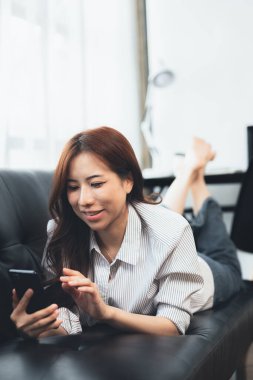 Asyalı bir kadın kanepede uzanıyor, bir arkadaşıyla akıllı telefondan uygulamayla mesajlaşıyor, yorucu bir iş gününden sonra hafta sonu molası veriyor. Tatil gevşeme fikirleri.