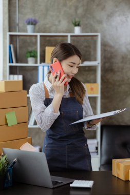 Asyalı bir kadın siparişi doğrulamak için bir müşteriyle telefonda konuşuyor, bir online mağazası var, özel bir nakliye şirketi aracılığıyla paketleyip gönderiyor. Çevrimiçi satış ve online alışveriş kavramları.