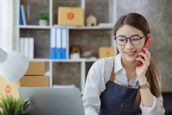 Asian woman is talking on the phone with a customer to confirm an order, she owns an online store, she packs and ships through a private transport company. Online selling and online shopping concepts.