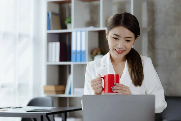 An Asian female employee sits with a red coffee mug in the startup\'s marketing department office, she is a marketing, customer liaison and consulting worker. Marketing concept.