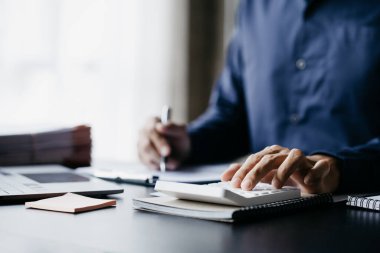 Businessman is using a calculator to calculate company financial figures from earnings papers, a businessman sitting in his office where the company financial chart is placed. Banner with copy space.