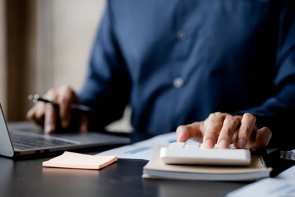 Businessman is using a calculator to calculate company financial figures from earnings papers, a businessman sitting in his office where the company financial chart is placed. Banner with copy space.