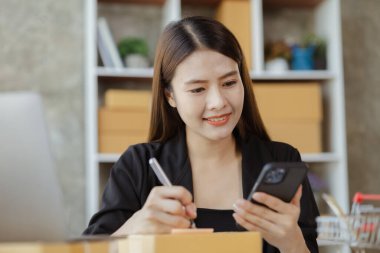 Asyalı bir kadın siparişi doğrulamak için bir müşteriyle telefonda konuşuyor, bir online mağazası var, özel bir nakliye şirketi aracılığıyla paketleyip gönderiyor. Çevrimiçi satış ve online alışveriş kavramları.