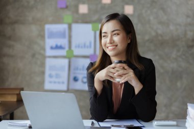 An Asian female employee sits with a coffee cup in the startup's marketing department office, she is a marketing, customer liaison and consulting worker. Marketing concept.