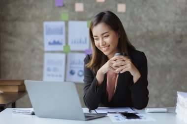 An Asian female employee sits with a coffee cup in the startup's marketing department office, she is a marketing, customer liaison and consulting worker. Marketing concept.