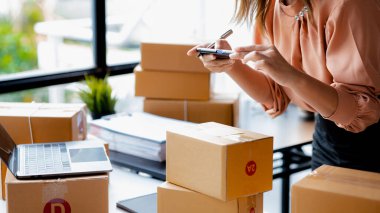 A woman using a smartphone to take pictures in front of parcel boxes, parcel boxes for packing goods, delivering goods through private courier companies. Online selling and online shopping concepts.