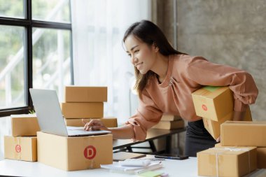 A beautiful Asian business owner opens an online store, she is checking orders from customers, sending goods through a courier company, concept of a woman opening an online business.