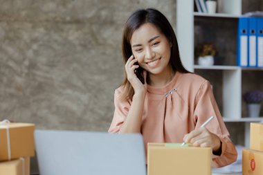 Asyalı bir kadın siparişi doğrulamak için bir müşteriyle telefonda konuşuyor, bir online mağazası var, özel bir nakliye şirketi aracılığıyla paketleyip gönderiyor. Çevrimiçi satış ve online alışveriş kavramları.