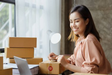 An Asian woman tying a parcel to a customer's box, she owns an online store, she packs and ships through a private transport company. Online selling and online shopping concepts.