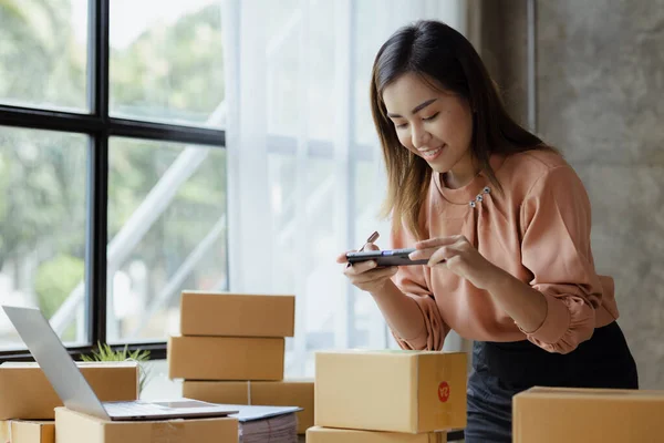 A woman using a smartphone to take pictures in front of parcel boxes, parcel boxes for packing goods, delivering goods through private courier companies. Online selling and online shopping concepts.