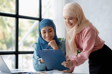 Two hijab-clad Asian women are brainstorming together in a conference room for a startup, run by a young, talented woman. The management concept runs the company of female leaders to grow the company.