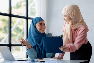 Two hijab-clad Asian women are brainstorming together in a conference room for a startup, run by a young, talented woman. The management concept runs the company of female leaders to grow the company.