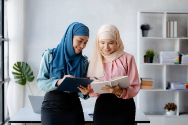 Two hijab-clad Asian women are brainstorming together in a conference room for a startup, run by a young, talented woman. The management concept runs the company of female leaders to grow the company.