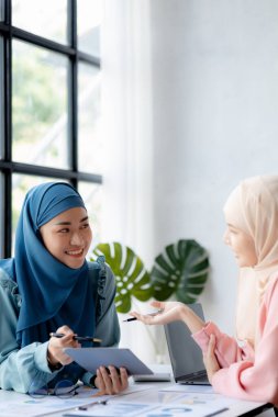 Two hijab-clad Asian women are brainstorming together in a conference room for a startup, run by a young, talented woman. The management concept runs the company of female leaders to grow the company.