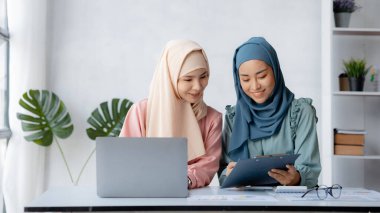 Two hijab-clad Asian women are brainstorming together in a conference room for a startup, run by a young, talented woman. The management concept runs the company of female leaders to grow the company.