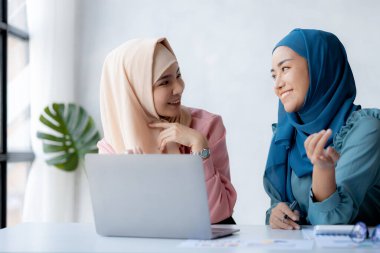 Two hijab-clad Asian women are brainstorming together in a conference room for a startup, run by a young, talented woman. The management concept runs the company of female leaders to grow the company.
