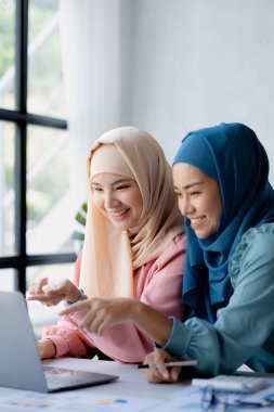 Two hijab-clad Asian women are brainstorming together in a conference room for a startup, run by a young, talented woman. The management concept runs the company of female leaders to grow the company.