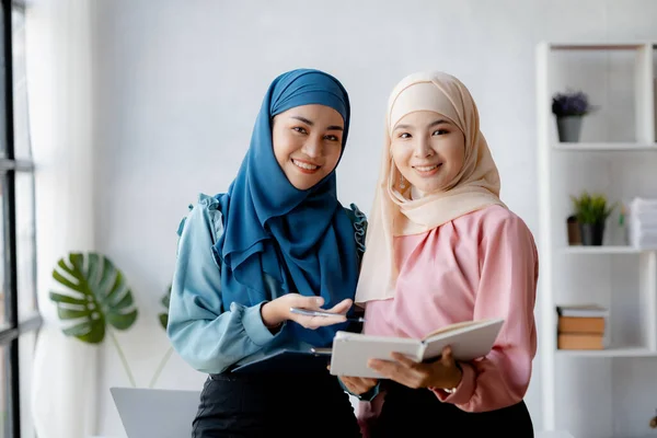 Two hijab-clad Asian women are brainstorming together in a conference room for a startup, run by a young, talented woman. The management concept runs the company of female leaders to grow the company.