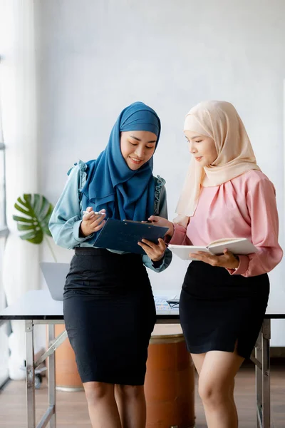 Two hijab-clad Asian women are brainstorming together in a conference room for a startup, run by a young, talented woman. The management concept runs the company of female leaders to grow the company.