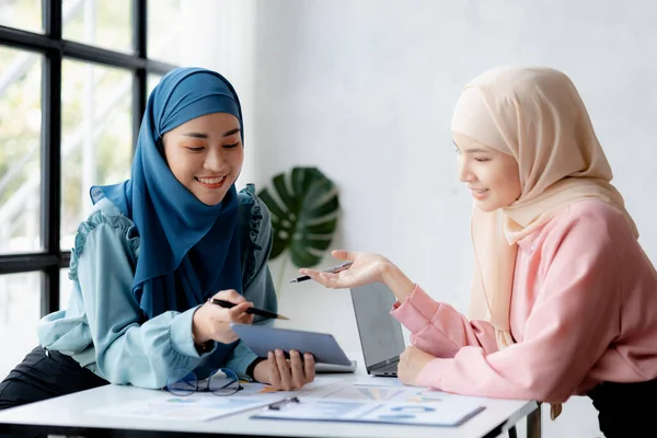 Two hijab-clad Asian women are brainstorming together in a conference room for a startup, run by a young, talented woman. The management concept runs the company of female leaders to grow the company.