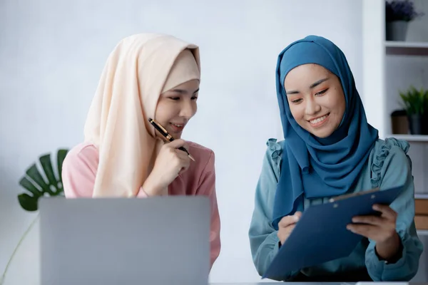 Two hijab-clad Asian women are brainstorming together in a conference room for a startup, run by a young, talented woman. The management concept runs the company of female leaders to grow the company.