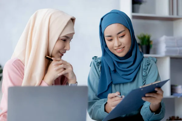 Two hijab-clad Asian women are brainstorming together in a conference room for a startup, run by a young, talented woman. The management concept runs the company of female leaders to grow the company.