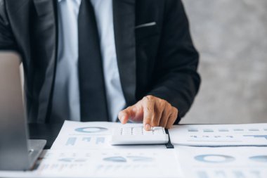 Businessman is using a calculator to calculate company financial figures from earnings papers, a businessman sitting in his office where the company financial chart is placed. Banner with copy space.