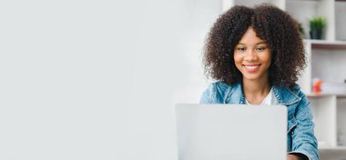 American teenage woman sitting in white office with laptop, she is a student studying online with laptop at home, university student studying online, online web education concept.