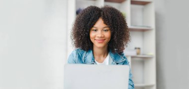American teenage woman sitting in white office with laptop, she is a student studying online with laptop at home, university student studying online, online web education concept.