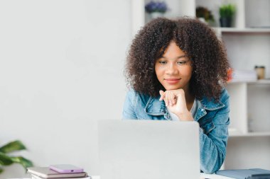 American teenage woman sitting in white office with laptop, she is a student studying online with laptop at home, university student studying online, online web education concept.