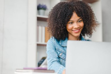 American teenage woman sitting in white office with laptop, she is a student studying online with laptop at home, university student studying online, online web education concept.